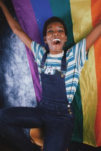 Image of a person smiling wide and holding a rainbow pride flag above their head. This image illustrates how freeing working with a Black queer therapist in Baltimore, MD for domestic violence counseling can be. | 21286 | 21044