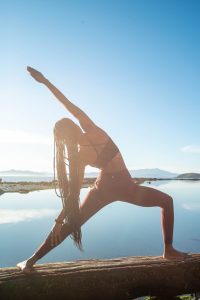 Image of a woman standing in front of a lake and practicing a yoga pose. This image represents what someone may look like practicing the mind-body medicine skill of a movement meditation learned from a holistic therapist in Baltimore, MD. 21204 | 21286 | 21044
