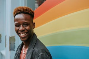 A young African American man stands in front of a wall painted in the colors of the rainbow representing the LGBTQ community. EMDR Therapy can help Black LGBTQ individuals overcome trauma.