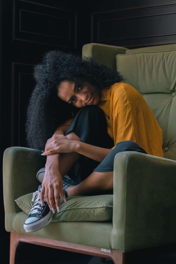 A woman in a gold shirt sits in a chair with her head resting on her knees as she smiles representing someone who has found relief from adrenal fatigue through Mind-Body counseling in Baltimore, MD. Contact a trauma therapist in Baltimore, MD to learn more about trauma healing in Baltimore, MD, and more. 